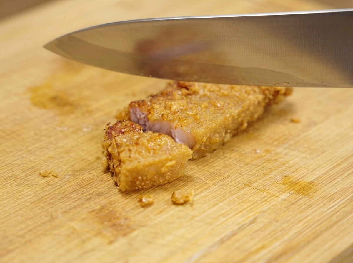 Ein großes Metallmesser schneidet ein knuspriges, goldbraunes Stück frittierten Schweinebauch auf einem Holzbrett in kleinere Portionen.