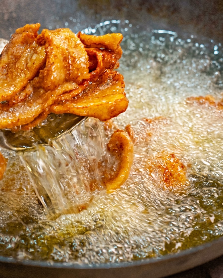 Goldbraune, gewellte Schweinebauchscheiben werden mit einer Schaumkelle aus einem Wok mit heißem Öl gehoben.