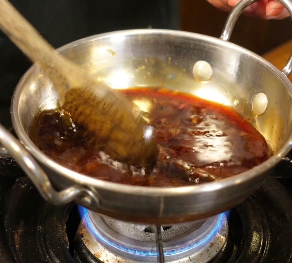 Ein Kochlöffel rührt eine dunkle, leicht köchelnde Sauce in einem kleinen Edelstahltopf über einer Gasflamme.