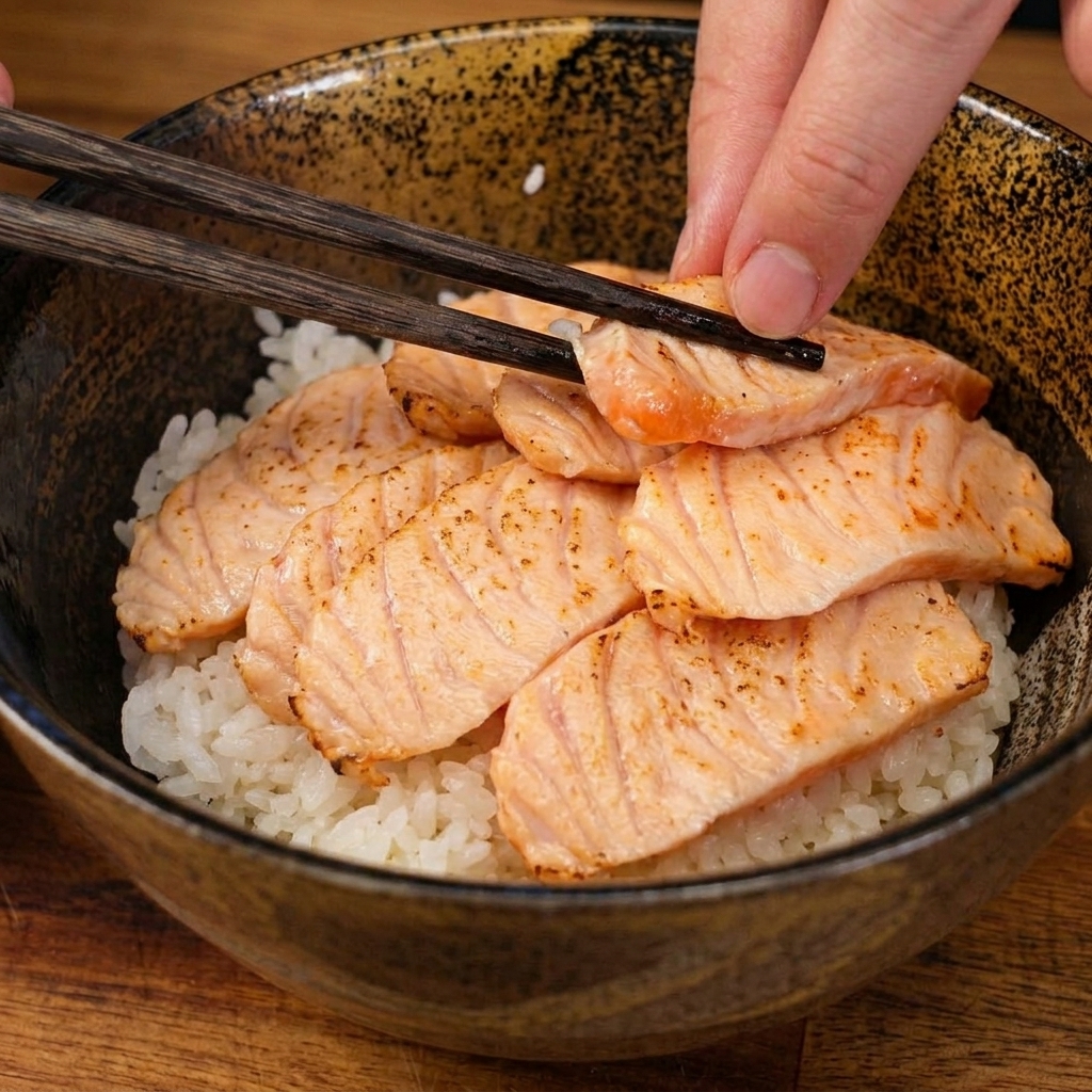 Essstäbchen legen eine Scheibe des kurz flambierten Lachs auf das Bett aus weißem Sushireis in einer Bowl.