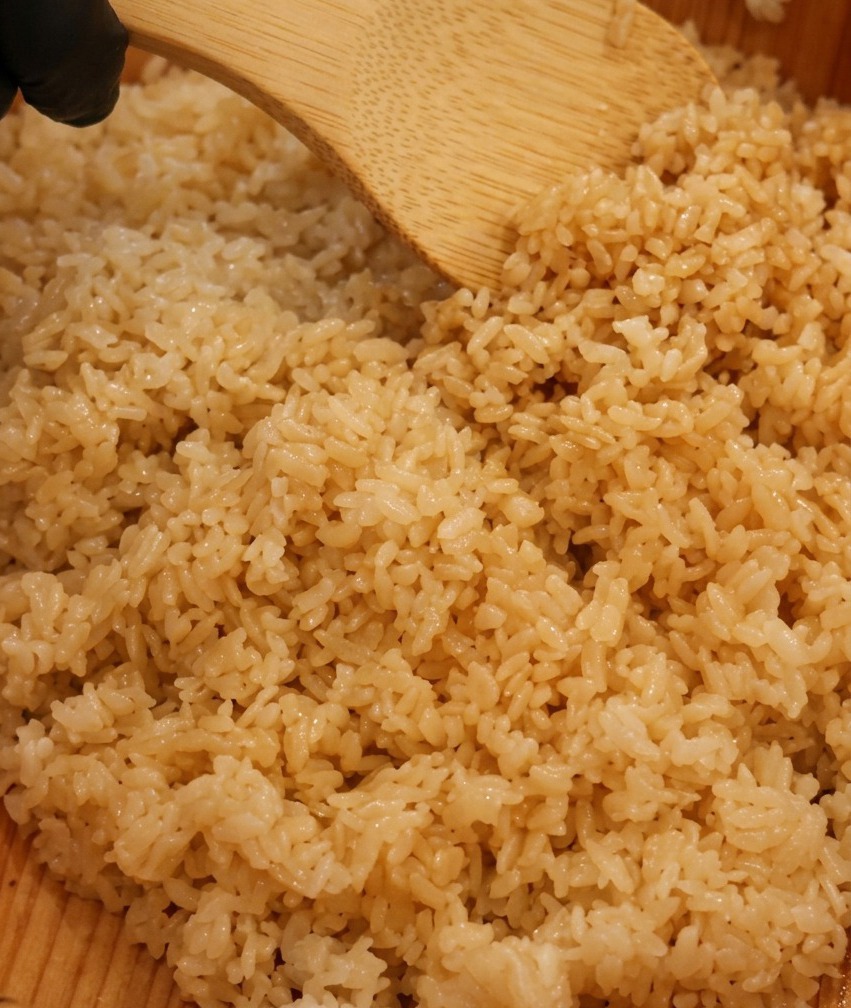 Dampfender Sushireis wird in einer Holzschale mit Sushieszig gewürzt und vorsichtig mit einem Holzspatel gemischt.