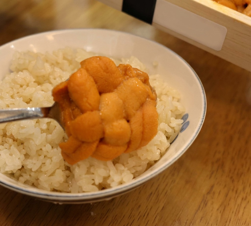 Ein Löffel hebt eine große, strukturierte Zunge von orangefarbenem Seeeigel an, um sie auf einer Schale mit weißem Sushireis zu platzieren.