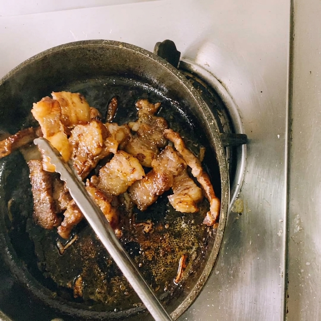 Eine Küchenzange aus Metall hält ein angebratenes Stück Rindfleisch, während es mit einer Schere direkt über der Pfanne in kleinere Stücke geschnitten wird.
