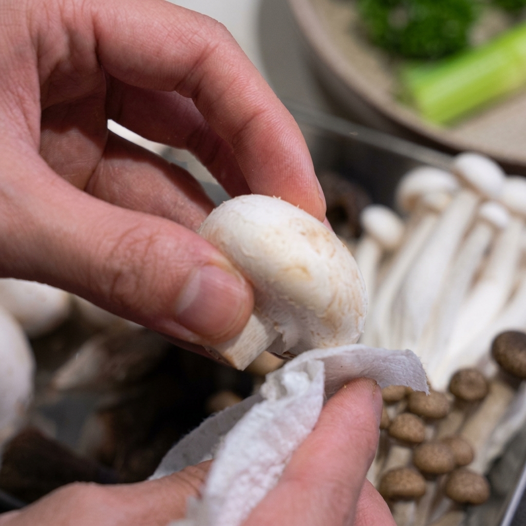 Nahaufnahme von Händen, die einen frischen weißen Champignon vorsichtig mit einem Küchentuch reinigen.