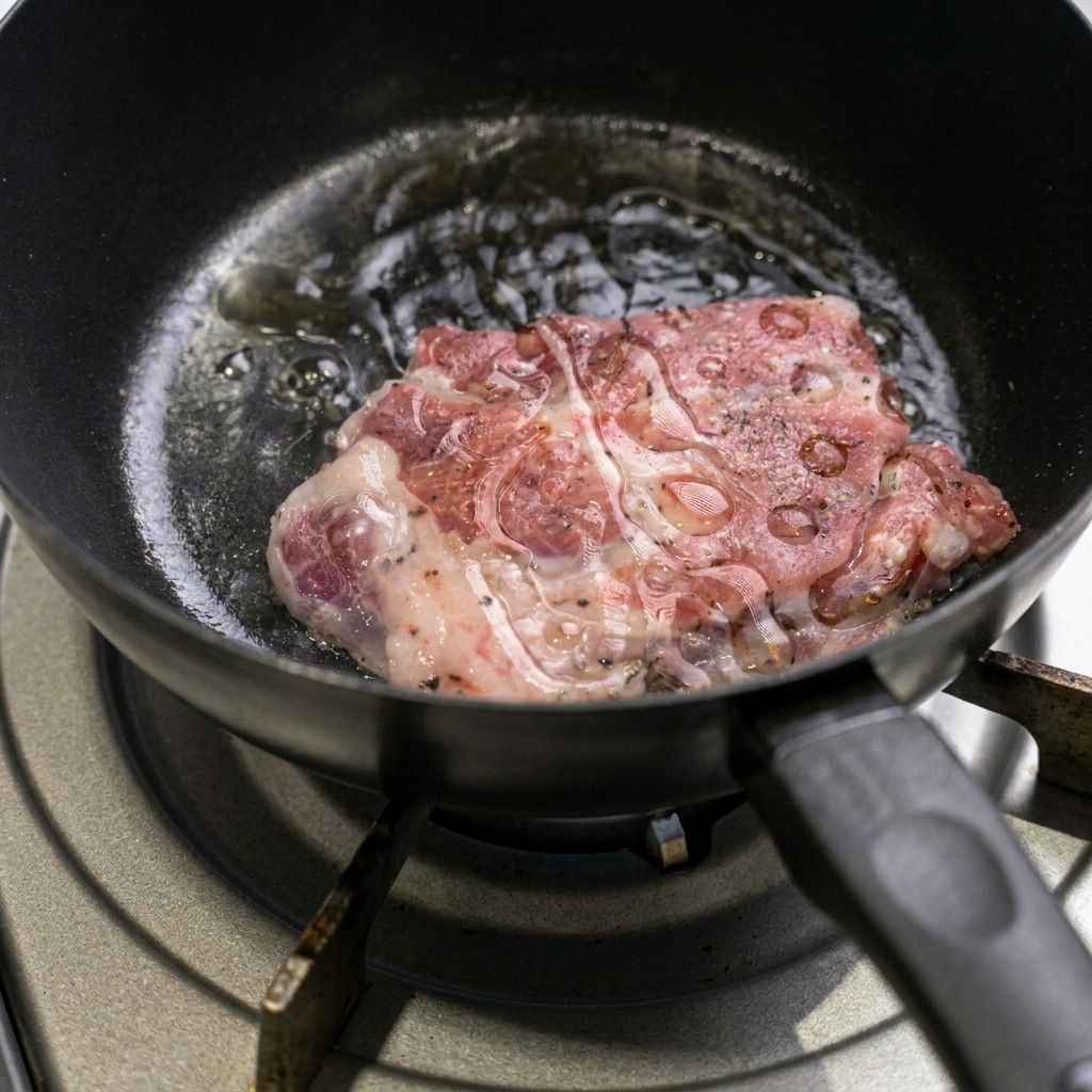 Ein gewürztes rohes Schweinenackensteak brät in einer heißen schwarzen Pfanne, während am Rand das Öl brodelt.