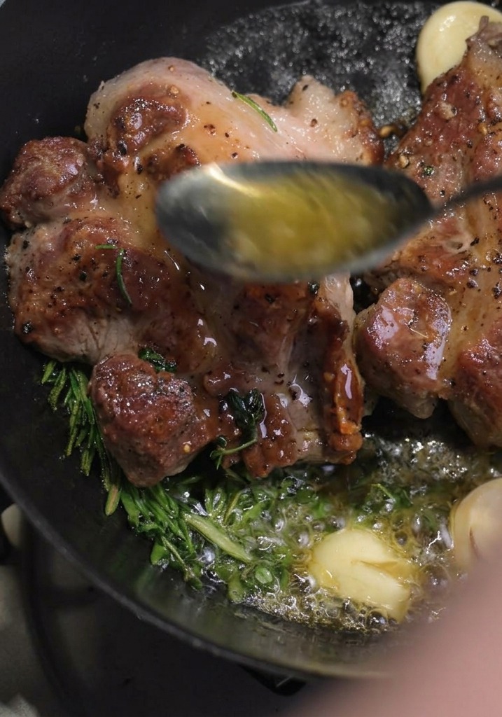 Ein Löffel schöpft geschmolzene heiße Butter auf die angebratenen Schweinenackensteaks, die mit Rosmarin und Knoblauch garen.
