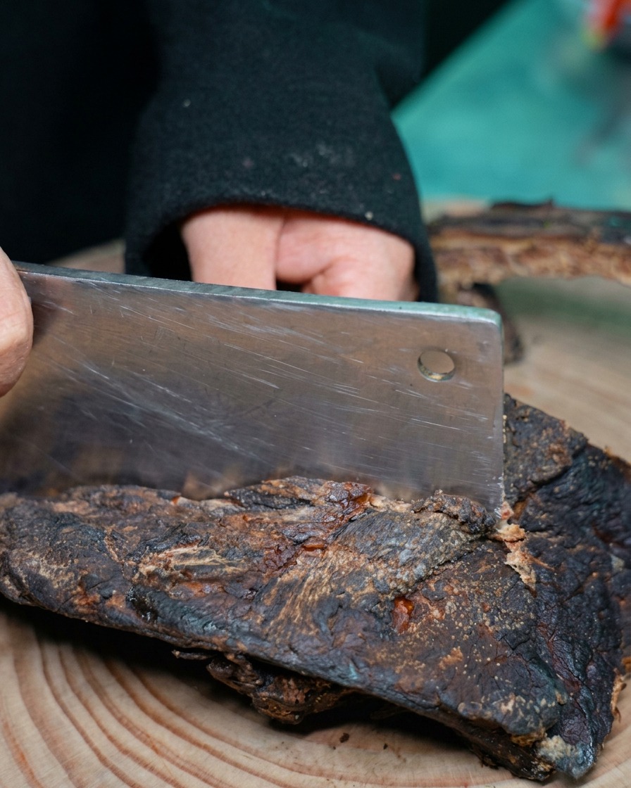 Eine Person hackt mit einem großen Hackmesser ein dunkles Stück gepökelte Schweinerippchen auf einem Holzschneidebrett.