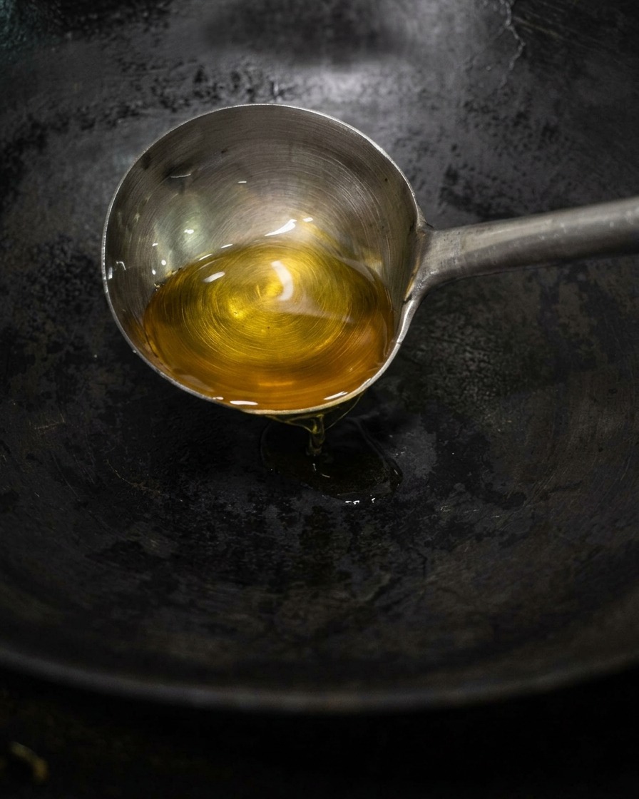Eine Metallkelle gießt goldenes Teeöl in einen dunklen, erhitzten Wok.