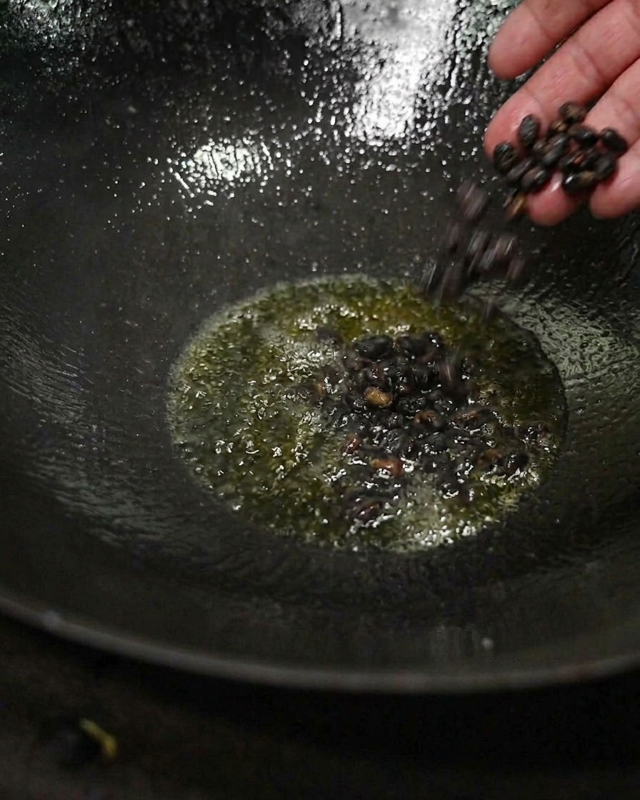 Eine Hand gibt eine kleine Menge dunkler, fermentierter schwarzer Bohnen in glänzend heißes Teeöl am Boden eines dunklen Woks.