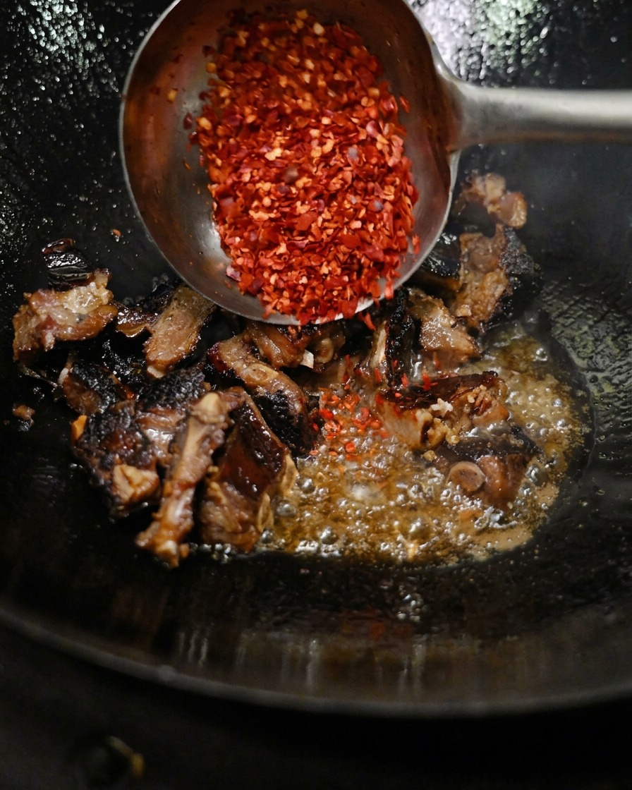 Eine Metallkelle streut gemahlenes rotes Chilipulver über gewürzte Schweinerippchen, die in einem heißen Wok gebraten werden.