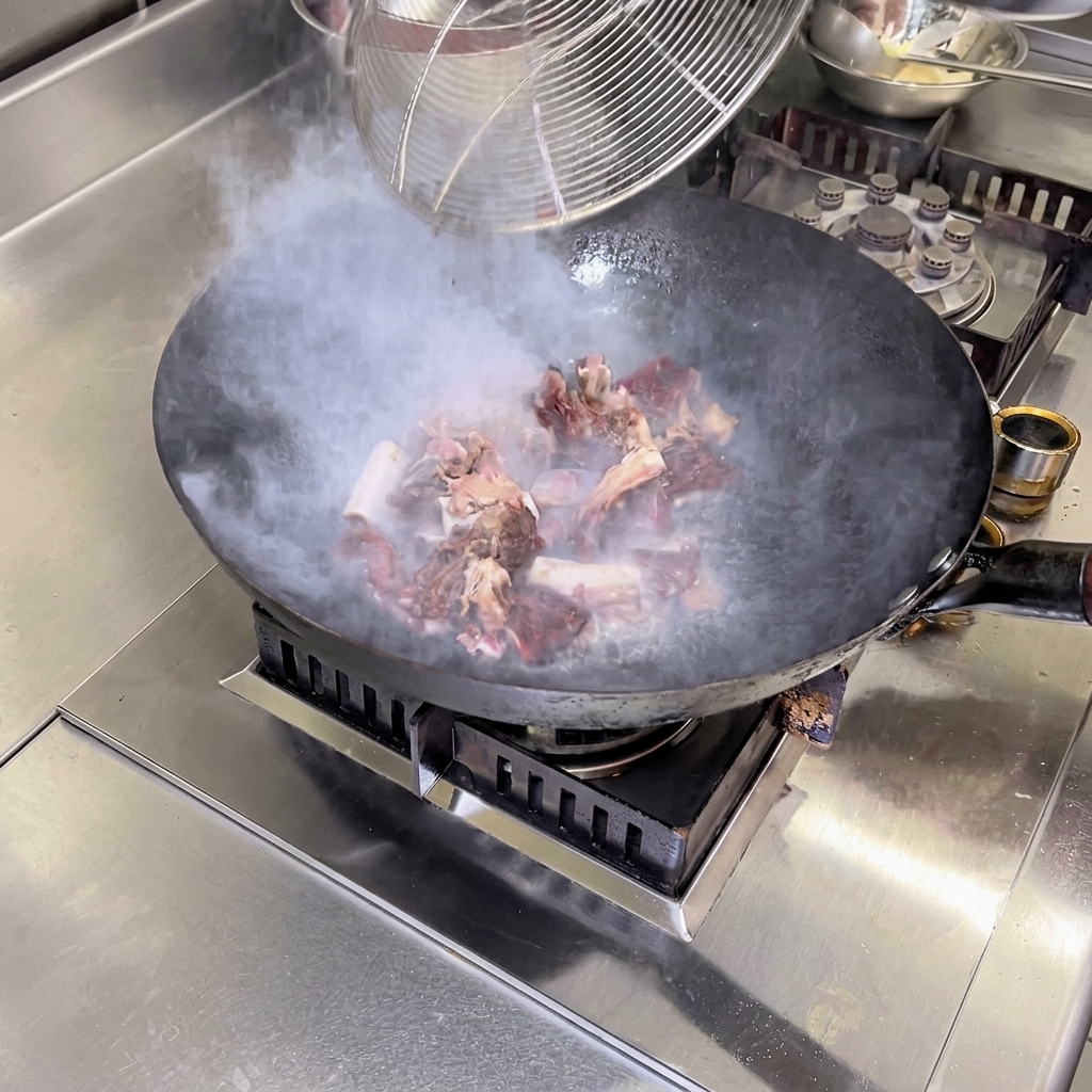Blanchierte gepökelte Schweinerippchen werden bei starker Hitze in einem heißen Wok mit Kamelienöl unter Rühren angebraten, während Dampf aus der Pfanne aufsteigt.