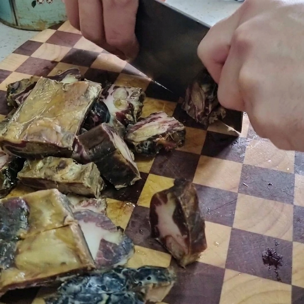 Nahaufnahme einer Person, die mit einem großen Hackmesser gepökelte Schweinerippchen auf einem hölzernen Schneidebrett in kleine Stücke schneidet.
