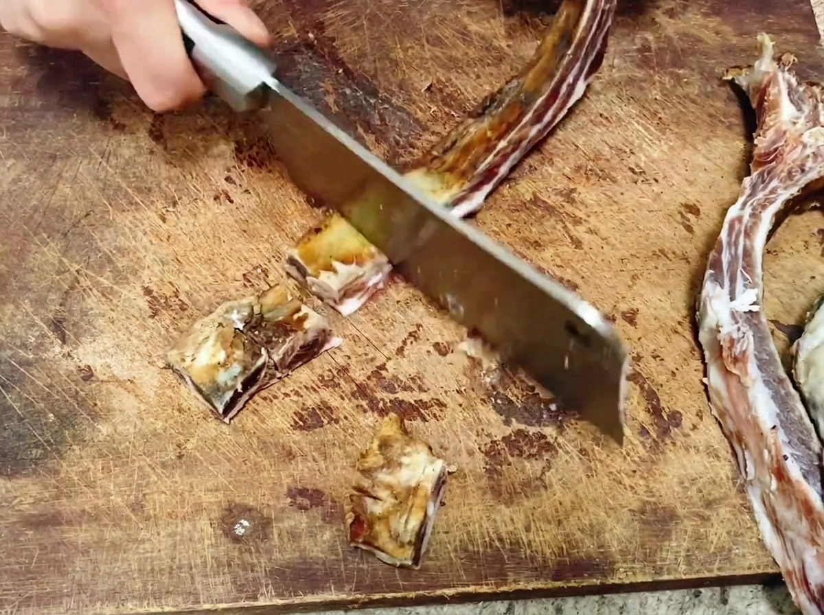 Ein großes Küchenbeil hackt eine lange gepökelte Schweinerippe auf einem Holzschneidebrett in mundgerechte Stücke.