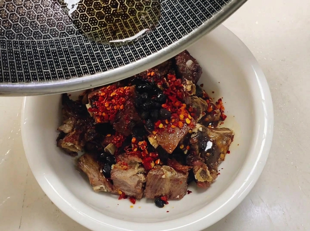 Warmes Kamelienöl wird aus einem Metallwok mit Wabenstruktur über eine gewürzte Schale mit Schweinerippchen, roten Chiliflocken und schwarzen Bohnen gegossen.