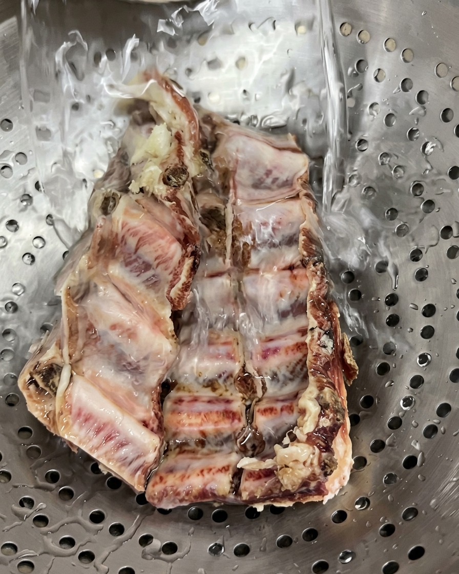 Rohe gepökelte Schweinerippchen in einem Edelstahlsieb werden unter fließendem kaltem Leitungswasser abgespült.