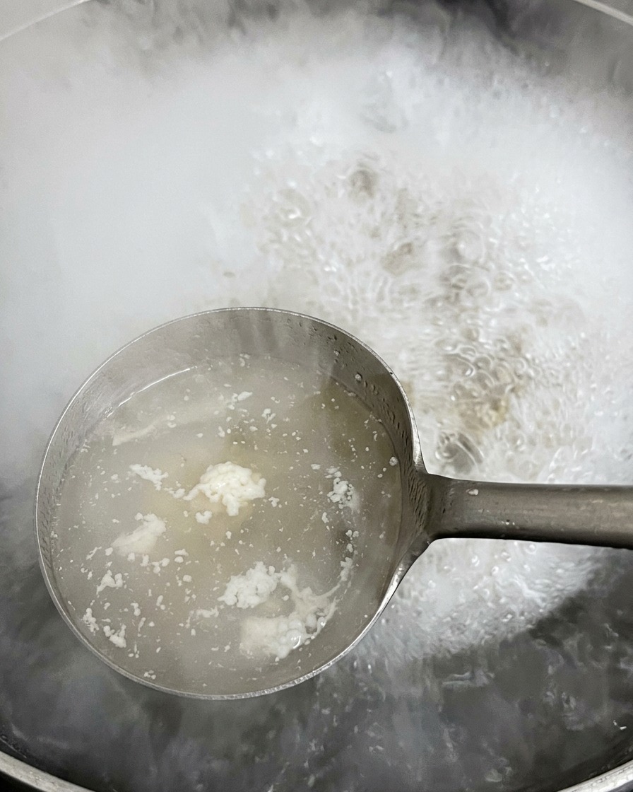 Eine Kelle aus Metall schöpft weißen, schäumenden Schmutz von der Oberfläche des kochenden Wassers in einem großen Topf.