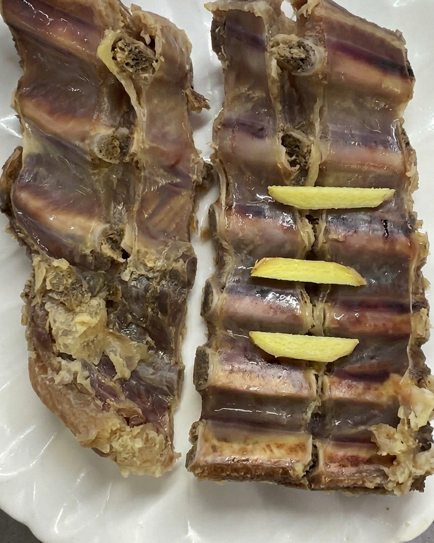 Zwei Stücke blanchierte Schweinerippchen liegen auf einem weißen Teller, ordentlich belegt mit frischen gelben Ingwerstreifen.