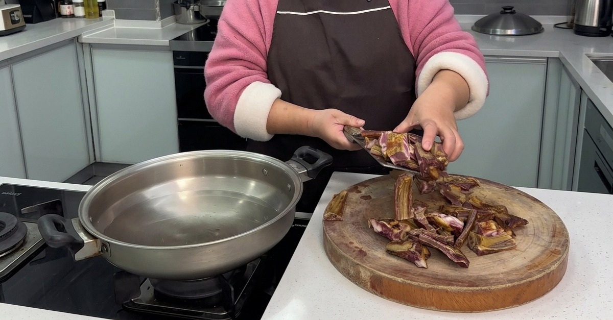 Eine Person gibt frisch gehackte Stücke von gepökelten Schweinerippchen von einem runden Holzschneidebrett in eine Edelstahlpfanne mit kaltem Wasser.