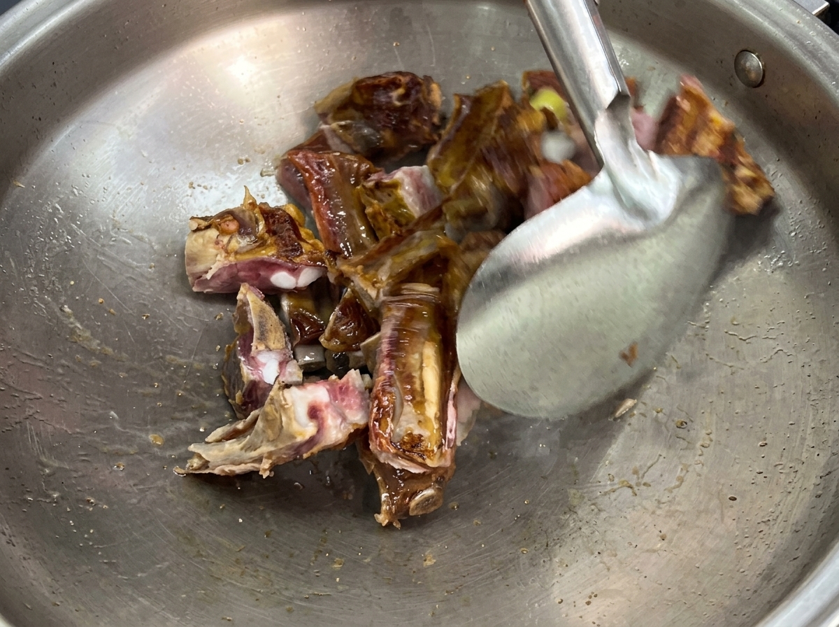 Ein Pfannenwender aus Metall brät gepökelte Schweinerippchen und in Scheiben geschnittenen Ingwer in einem großen Edelstahl-Wok an.