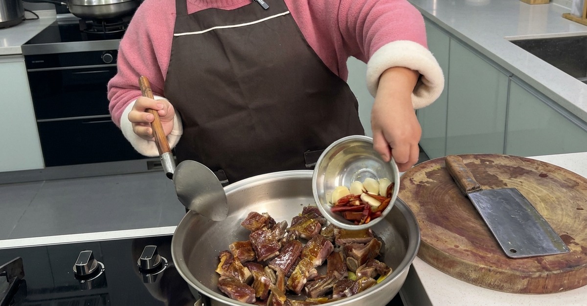 Eine Person schüttet ganze Knoblauchzehen und getrocknete rote Chilis aus einer kleinen Metallschüssel in einen Wok zu den angebratenen Rippchen.
