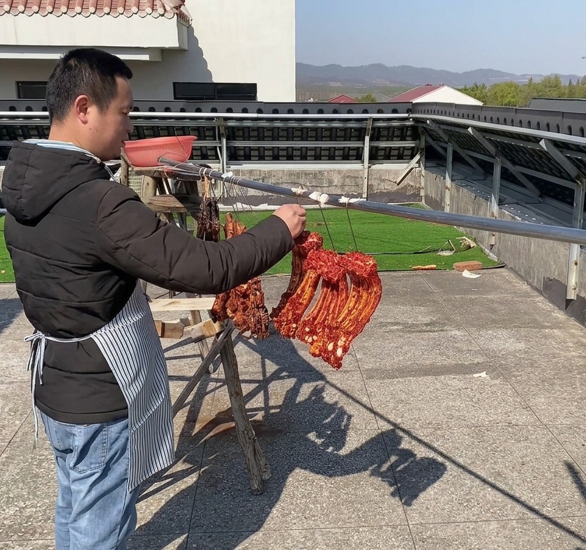 Kräftig gewürzte, tiefrote Schweinerippchen hängen nebeneinander an einer langen Metallstange im Freien, um unter klarem Himmel an der Luft zu trocknen.
