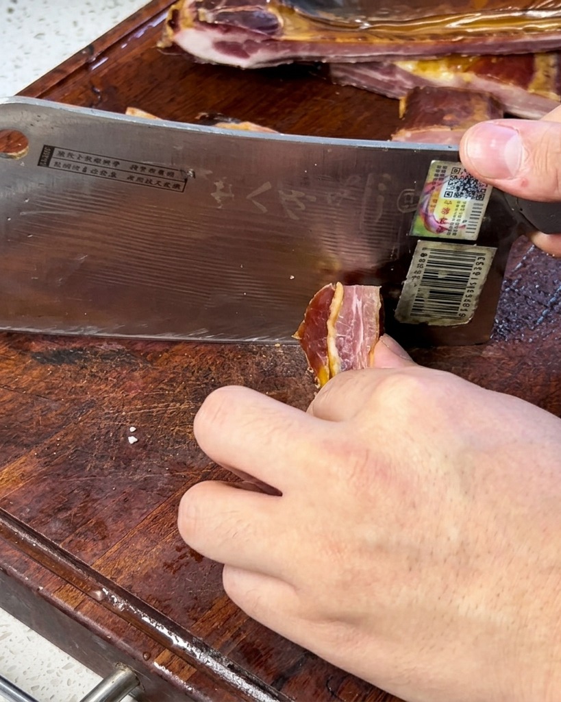 Eine Nahaufnahme eines Kochs, der mit einem breiten Küchenbeil dünne Scheiben von gepökeltem Schweinefleisch auf einem Holzschneidebrett schneidet.