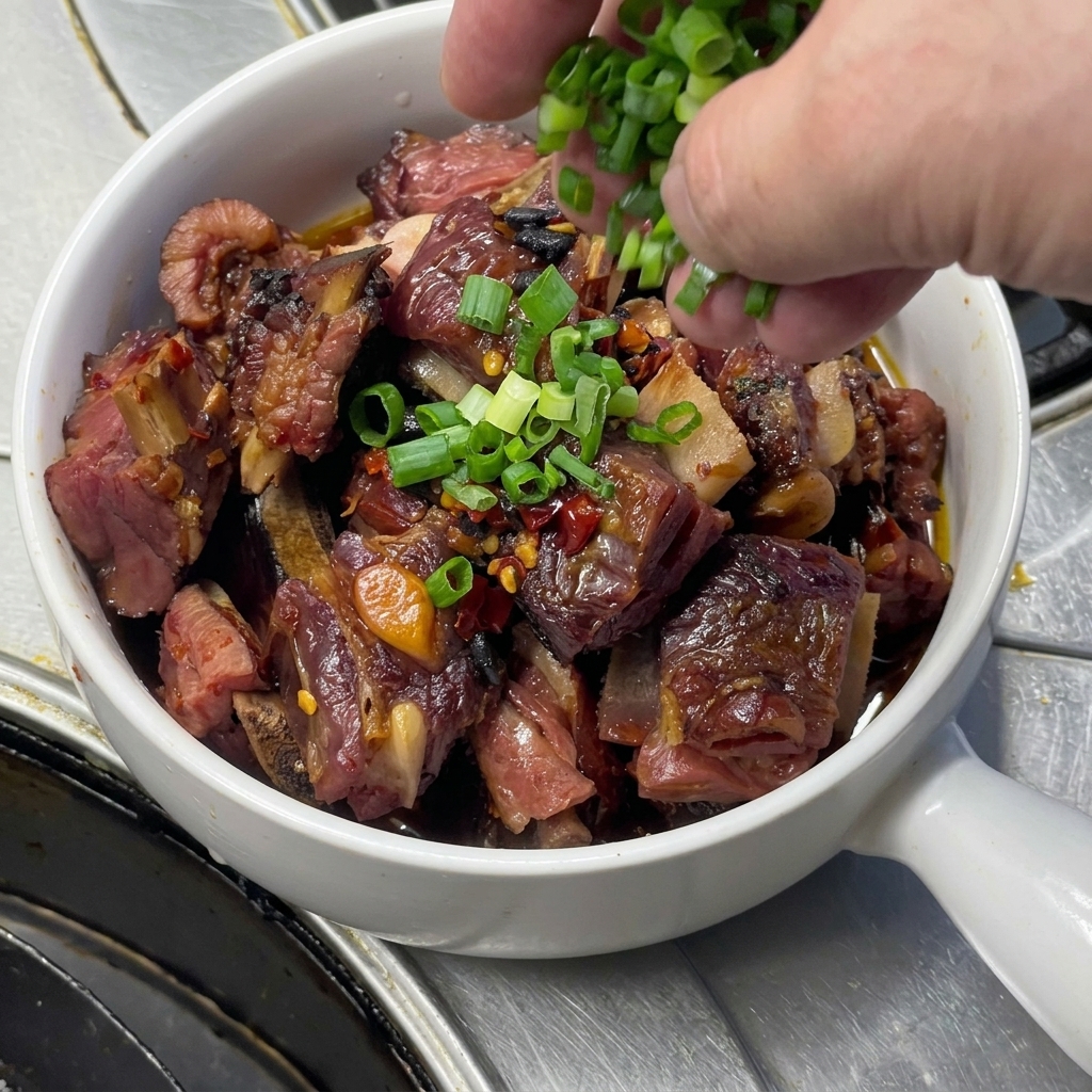 Frisch gehackte Frühlingszwiebeln werden aus einer Hand auf eine weiße Schüssel mit Henkel gestreut, die mit glänzenden gedämpften Schweinerippchen, Knoblauchzehen und Chili gefüllt ist.
