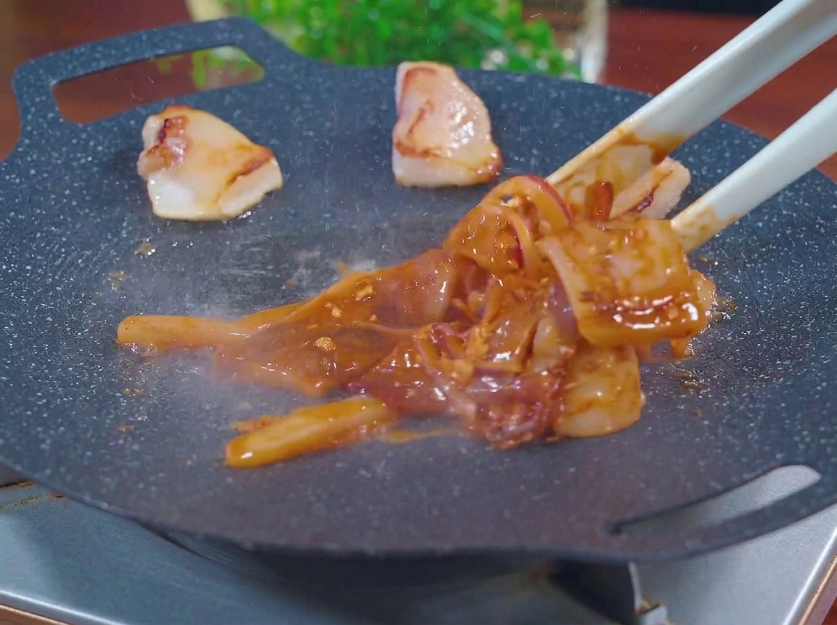 Eine Zange legt marinierten roten Tintenfisch und Zwiebelstreifen in eine heiße, dampfende schwarze Grillpfanne neben ausgelassene Speckstücke.