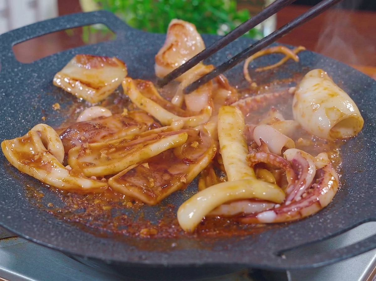Tintenfischstücke werden mit Essstäbchen auf einer flachen, heißen Grillplatte gewendet und brutzeln in einer dicken, blubbernden rötlich-braunen Barbecue-Marinade, während Dampf aufsteigt.