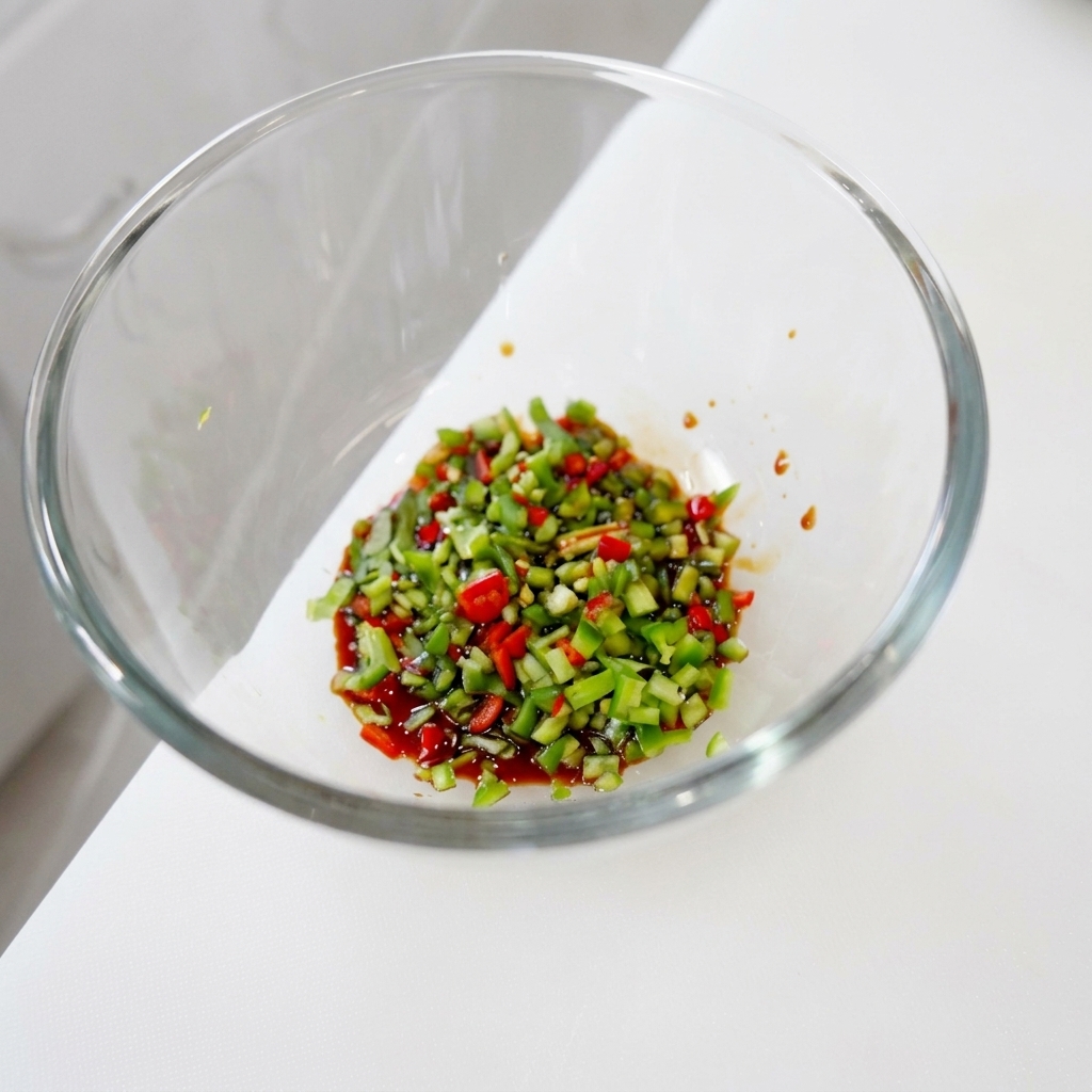 Eine bunte Mischung aus fein gehackten grünen und roten Chilischoten sowie Koriander zieht in einer dunklen, glänzenden flüssigen Gewürzbasis in einer Glasschüssel.