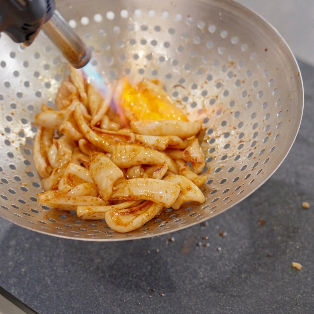 Frittierte Tintenfischstücke in einem großen, gelochten Metallsieb werden mit einer blau-orangefarbenen Flamme aus einem handgehaltenen Küchenbrenner abgeflämmt.