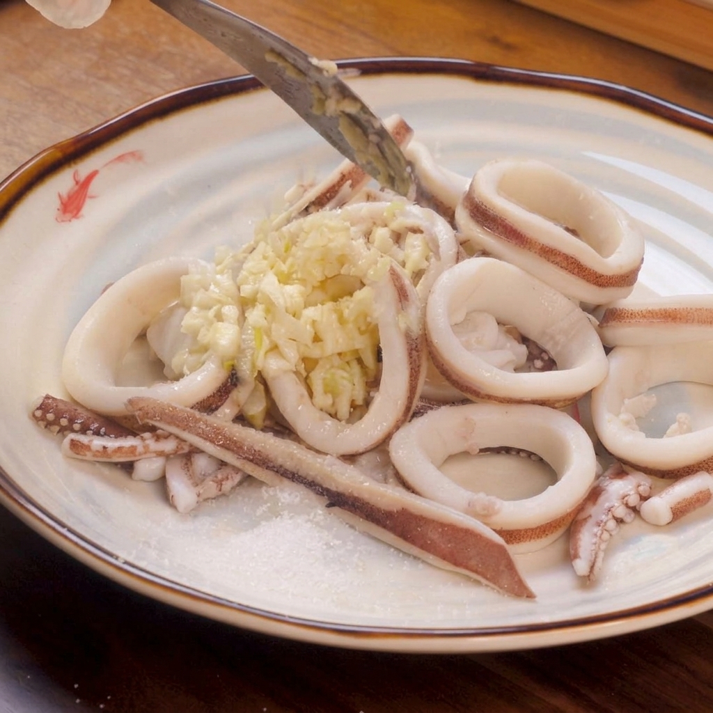 Blanchierte weiße Tintenfischringe auf einem Teller, garniert mit einer großzügigen Portion gehacktem Knoblauch sowie etwas Salz und Zucker am Rand.