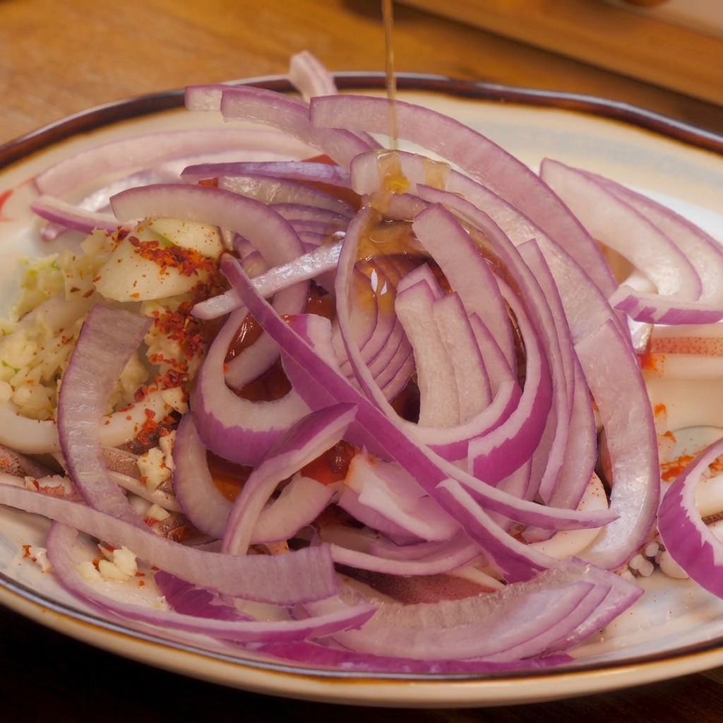 Ein feiner Strahl klares Sesamöl wird von oben über die bunten Zwiebelstreifen, das Chilipulver und den Tintenfisch gegossen.