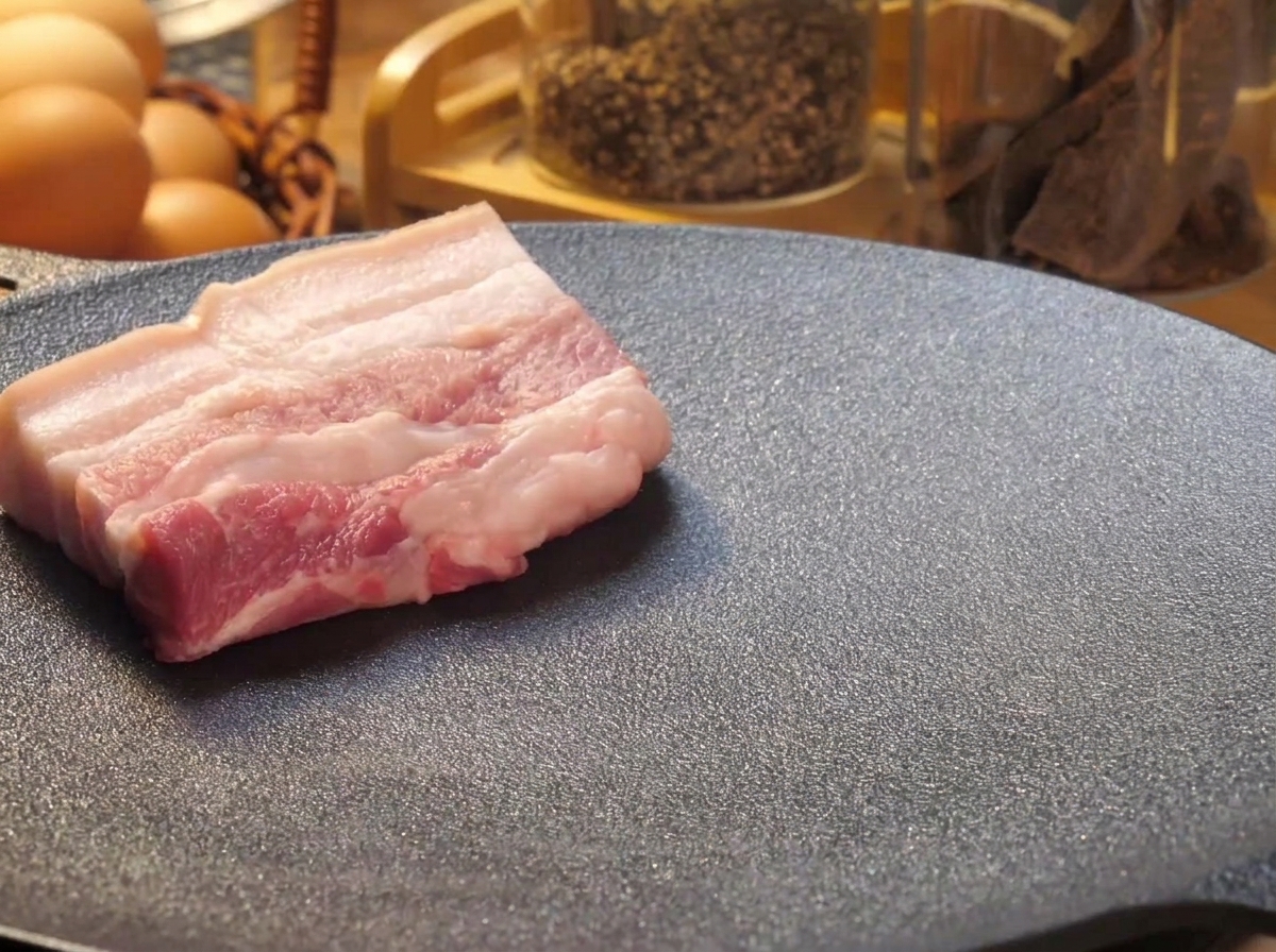 Dickere Scheiben roher Schweinebauch werden zum Anbraten auf eine heiße, schwarze Grillplatte gelegt.