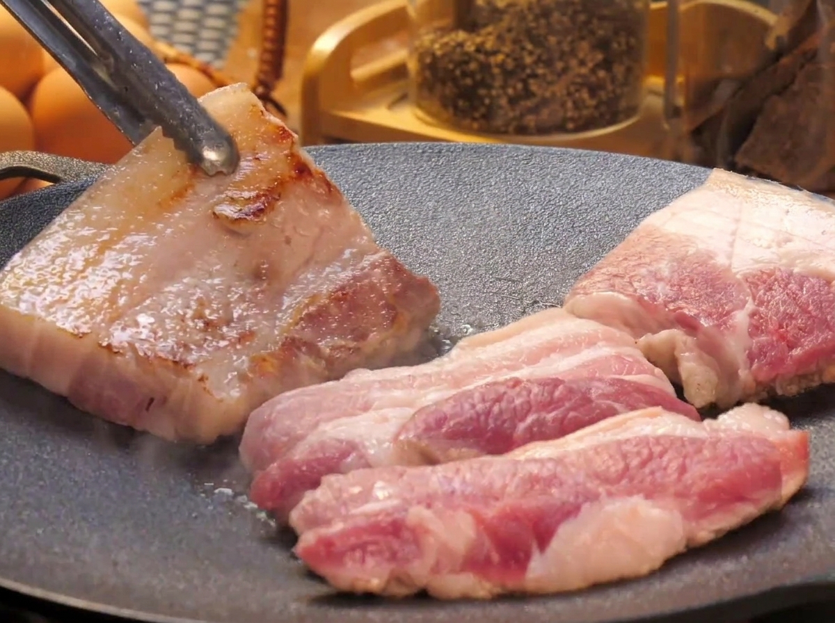 Schweinebauchscheiben brutzeln auf einer flachen Grillplatte; ein Stück wird mit einer Metallzange umgedreht und zeigt eine goldbraun krosse Oberfläche.