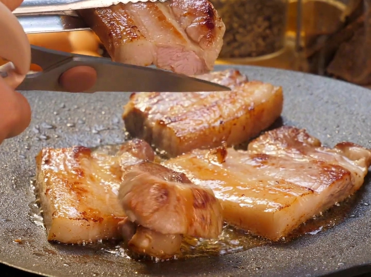 Eine Hand schneidet mit einer Küchenschere die dicken, krossen Schweinebauchscheiben direkt über der heißen Grillplatte in kleinere Streifen.