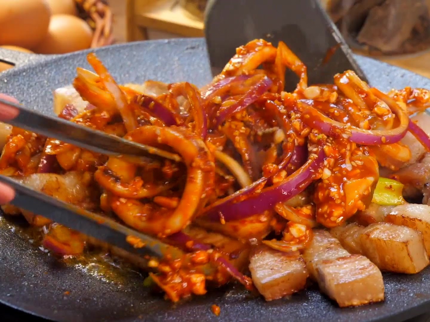Mit einem Pfannenwender und einer Zange werden die marinierten Tintenfischringe, Schweinebauchstreifen und roten Zwiebeln auf der brutzelnden Grillplatte miteinander verrührt.