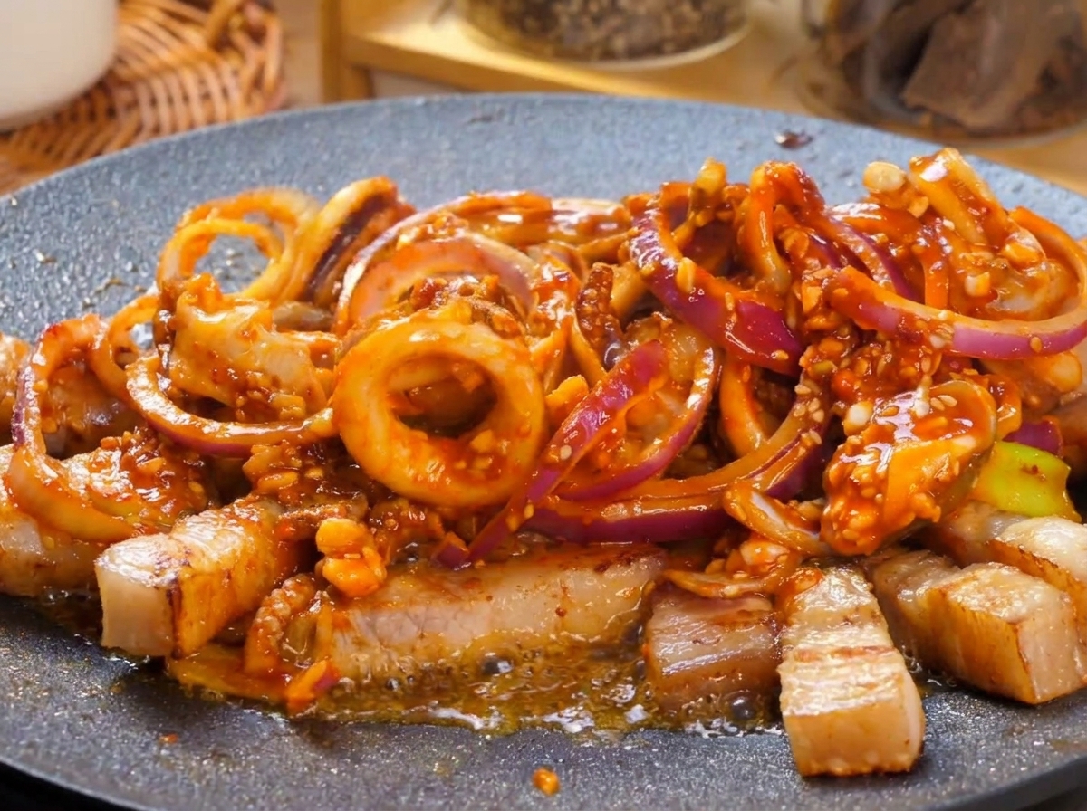 Eine heiße Grillplatte mit einem scharf brutzelnden Tintenfisch-Schweinebauch-Gericht in roher Sauce, garniert mit frischen grünen Chilischeiben.