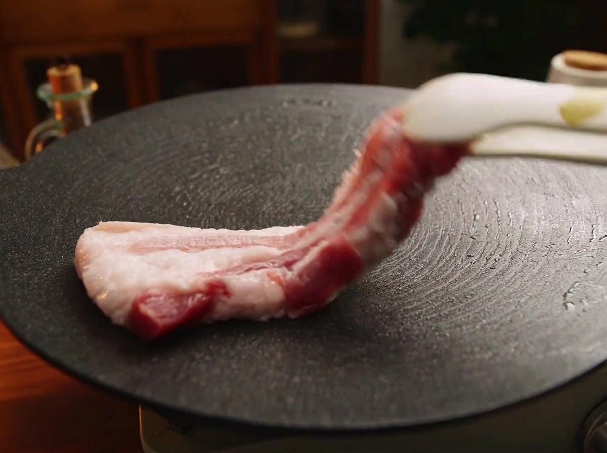Ein dicker Streifen roher Schweinebauch wird mit einer Zange auf eine heiße, schwarze Gusseisenplatte gelegt.
