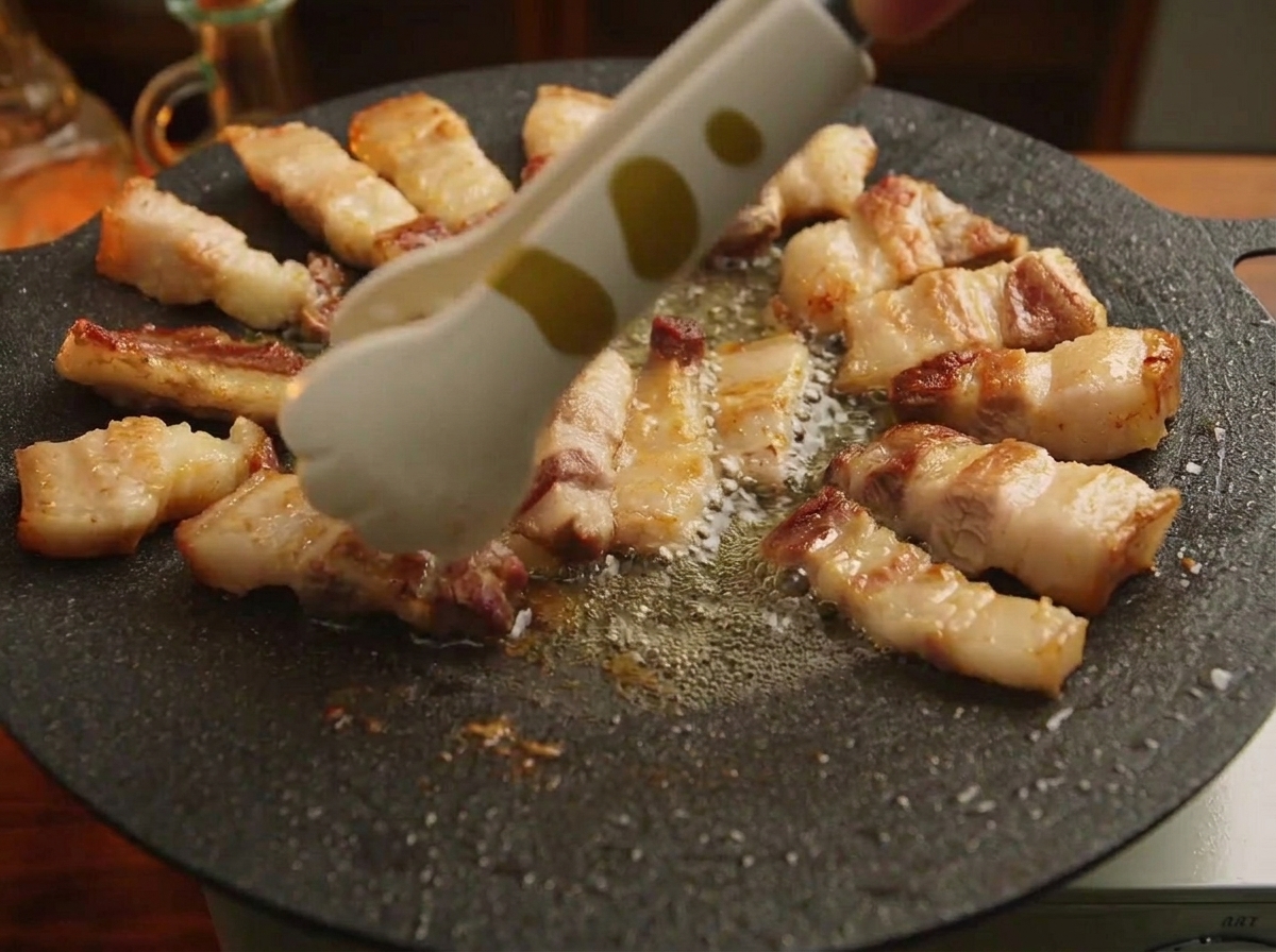 Eine Zange ordnet mundgerechte Stücke vom gegrillten Schweinebauch kreisförmig am Rand einer schwarzen Gusseisenplatte an.