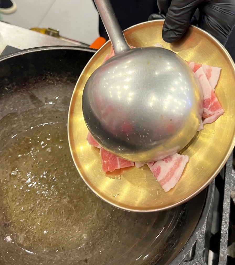 Rohe Schweinebauchscheiben werden mit einer großen Metallkelle von einem goldenen Teller in einen geölten Wok gegeben.