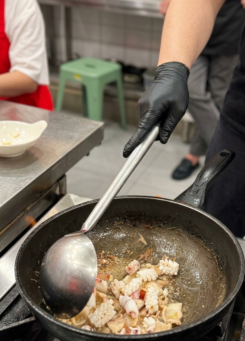 Nahaufnahme von eingeritzten Tintenfischstücken, die mit einer großen Metallkelle zusammen mit gebratenen Schweinebauchscheiben im Wok vermengt werden.