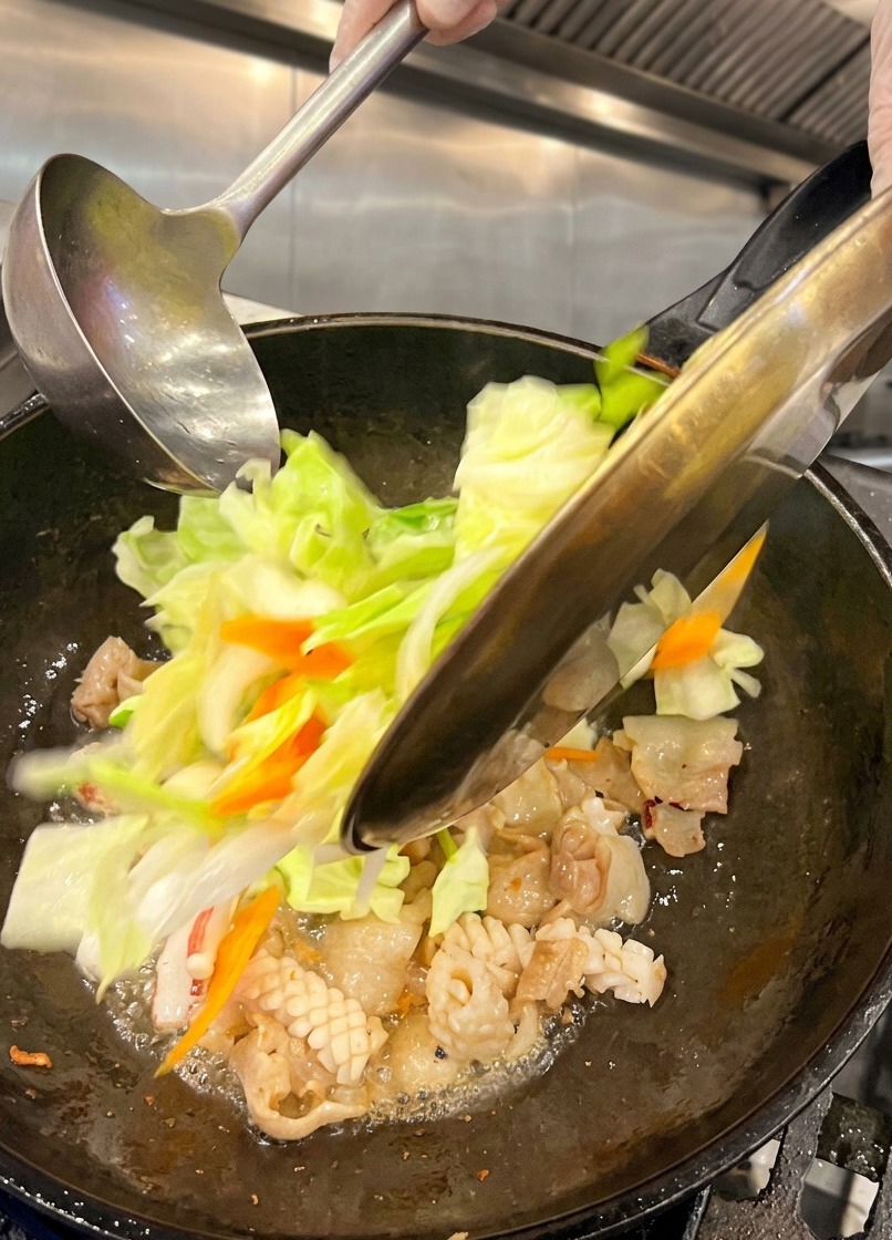 Frisch geschnittener Weißkohl und Karottenscheiben werden von einem Metallteller in einen heißen Wok mit Schweinebauch und Tintenfisch geschüttet.