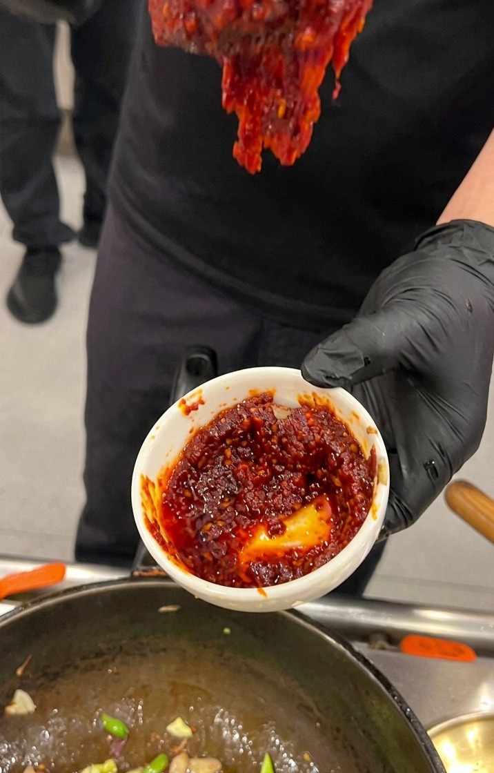 Eine Hand in einem schwarzen Handschuh hält eine kleine weiße Schale mit einer dickflüssigen, tiefroten koreanischen Chilipaste-Sauce über einen Wok.