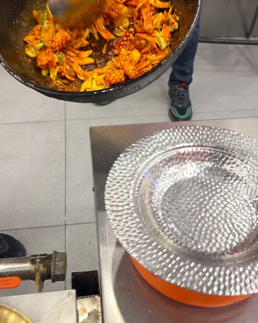 Pfannengerührter scharfer koreanischer Tintenfisch und Schweinebauch mit Gemüse werden mit einer Kelle aus dem Wok auf einen gehämmerten Silberteller gegeben.