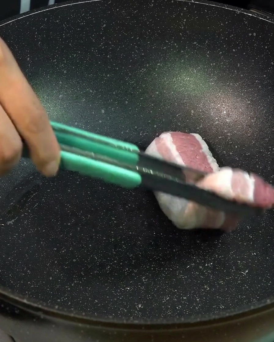 Dicke Scheiben roher Schweinebauch werden mit einer hellgrünen Zange zum Anbraten in eine schwarze Pfanne gelegt.