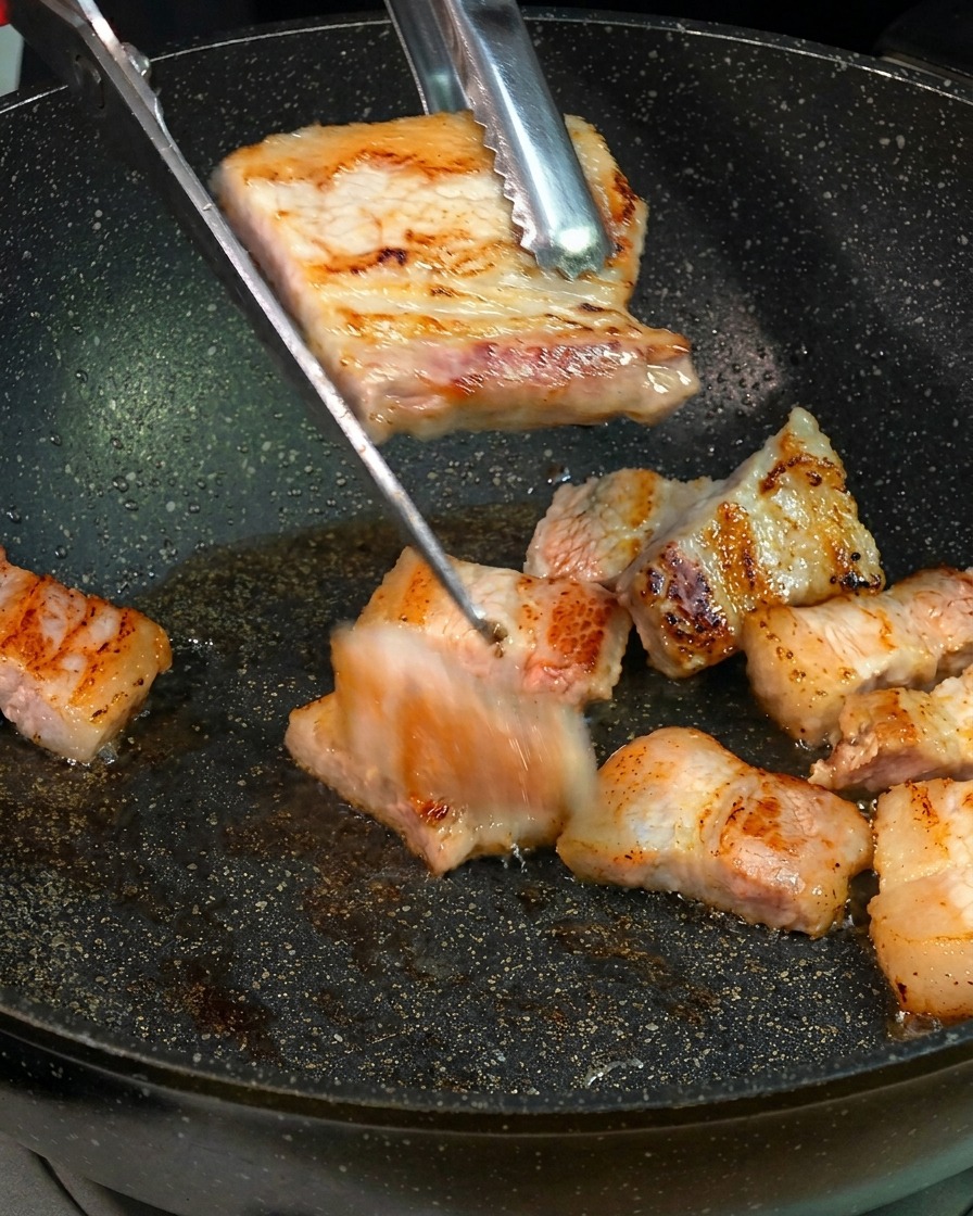 Goldbraun angebratene Schweinebauchscheiben werden mit einer Küchenschere direkt über einer Bratpfanne in kleinere Stücke geschnitten.