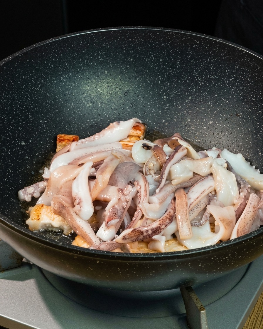 Frische Tintenfischstücke liegen über angebratenen Schweinebauchstücken in einem tiefen schwarzen Wok.