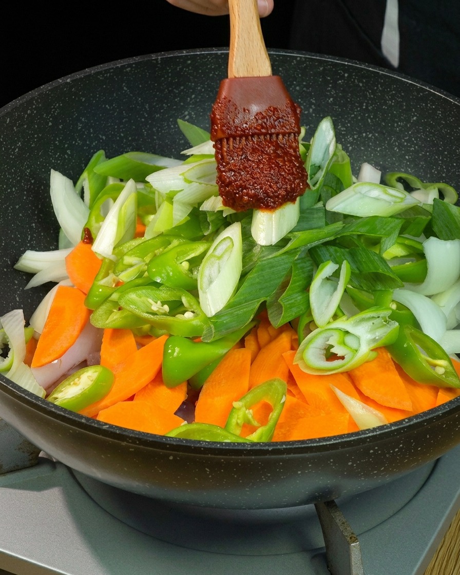 Ein dicker Klecks dunkelroter Chilisauce auf einem Holzspatel wird über frisch geschnittenen Karotten, grünen Chilis, Zwiebeln und Frühlingszwiebeln in einer Pfanne platziert.