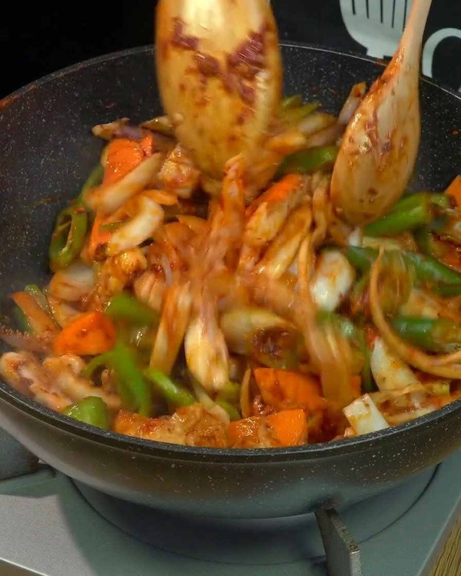 Bewegungsunschärfe von Schweinebauch, Tintenfisch und gemischtem Gemüse, die mit zwei Holzlöffeln kräftig in einem Wok geschwenkt und gebraten werden, vollständig mit roter scharfer Sauce überzogen.